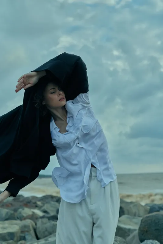 Fashion, wind, cloak on beach, woman in light shirt and loose pants under a cloudy sky, dramatic movement as she lifts a dark cloak, coastal scenery with sea and rocks