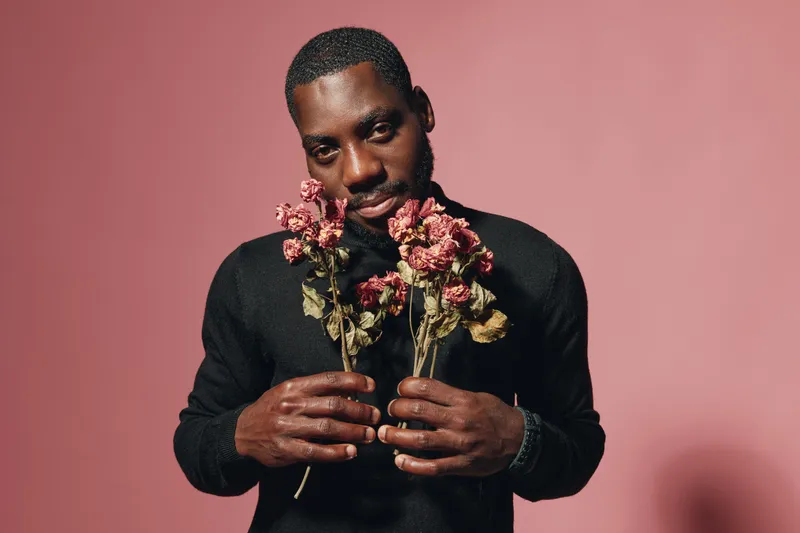 African man holding dried flowers in hands with creative conceptual visual style on pink background. Portrait of black male with artistic expression and fashion in studio.