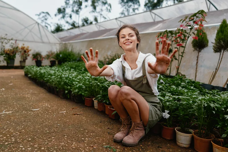 Smiling young woman in a greenhouse, showcasing healthy plants and embracing nature with joy and enthusiasm, dressed in casual attire, earthy color palette, vibrant atmosphere