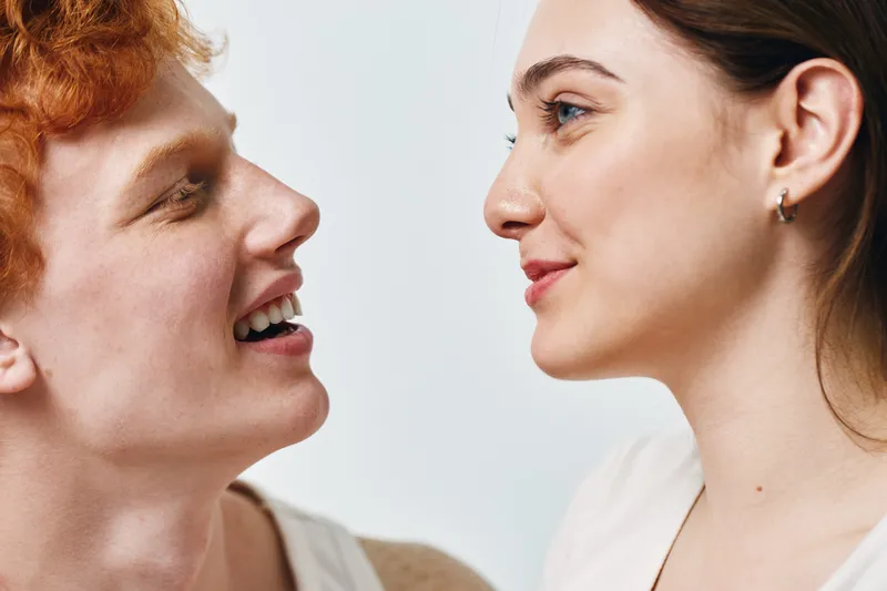 Two young people facing each other smiling with clear skin and natural makeup, closeup portrait showing connection and positive emotions indoors.