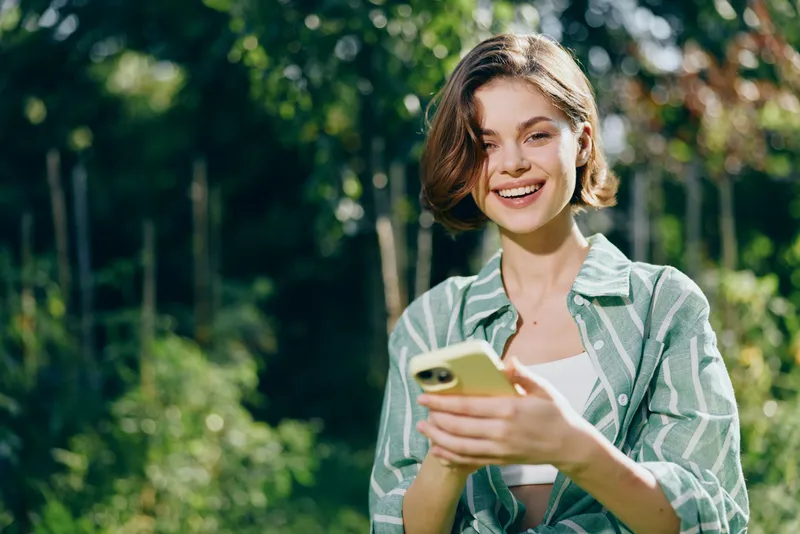 Casual young woman outdoors, smiling while using a smartphone in a sunlit park, waist up portrait with greenery background.