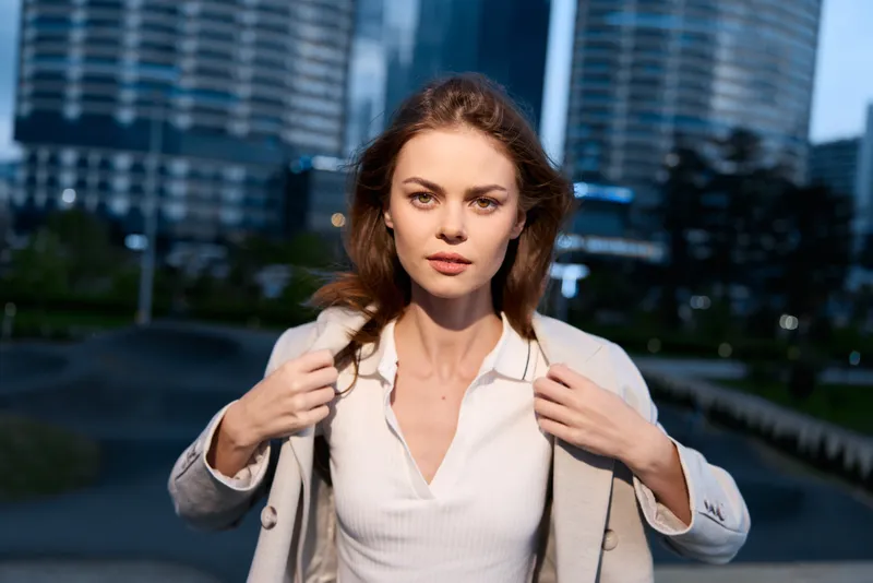 Confident woman in business attire poses outdoors with a city skyline background, showcasing her professionalism and determination in a modern corporate environment.