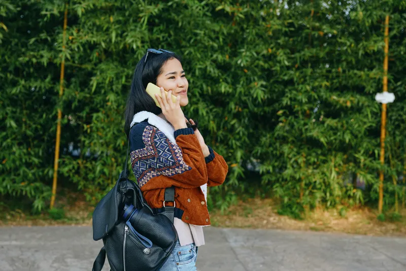 Woman on a casual outdoor walk talks on a smartphone, carrying a backpack beside a green hedge and street setting, illustrating mobile communication, travel lifestyle, and everyday errands