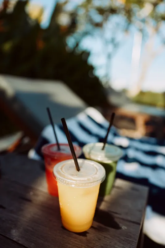 A trio of cold beverages on a wooden table outdoors in bright sun, straws upright. A relaxed lifestyle moment for casual gatherings, summer afternoons, and refreshing drinks in a carefree outdoor