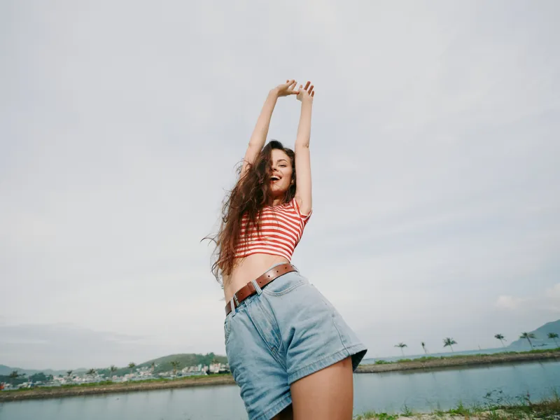 Joyful young woman outdoors with arms raised, wearing a striped crop top and denim shorts, smiling confidently under bright daylight against an expansive sky.