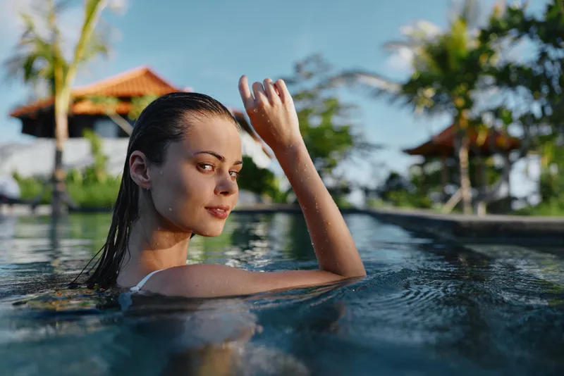 Young woman relaxing in a pool at a tropical resort, sunlit water and palm trees in the background, showcasing leisure, wellness, and vacation vibes.