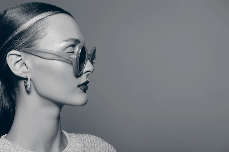Profile of a young white woman with pale skin wearing large sunglasses and hoop earrings in a minimalist setting.