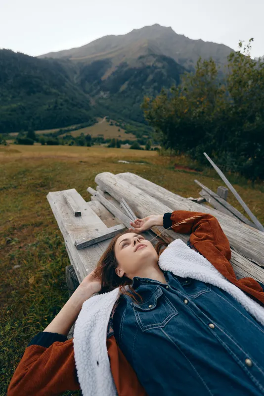 woman outdoors in meadow and mountains lying on wood planks, portrait of relaxation under open sky, casual denim jacket and cozy coat in peaceful natural landscape