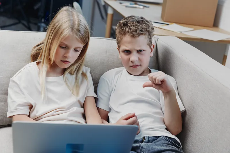 Two children sitting on a couch using a laptop with serious and displeased expressions. Concept of childhood technology use, frustration, and digital interaction.