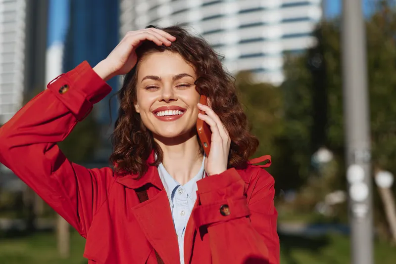 Young woman with curly hair smiles while talking on a phone outdoors in a city park, wearing a red jacket under sunny sky.
