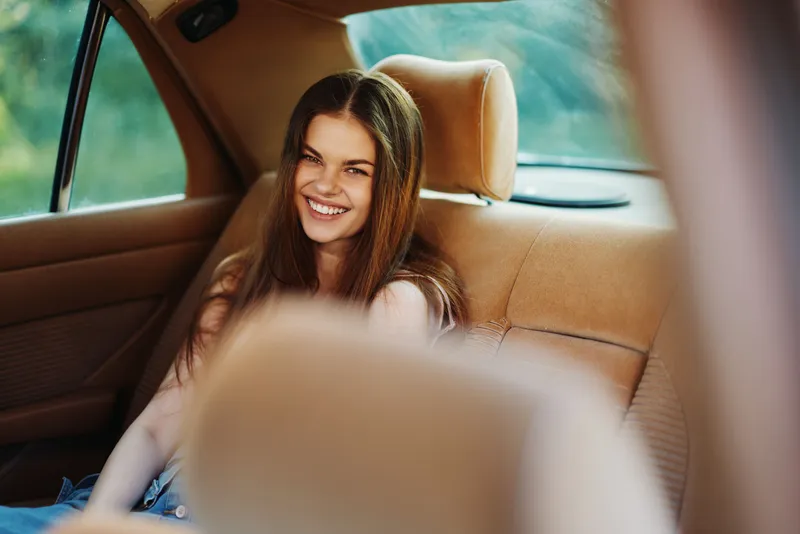 Young woman smiling joyfully in the backseat of a car, enjoying a sunny day with vibrant green foliage in the background.