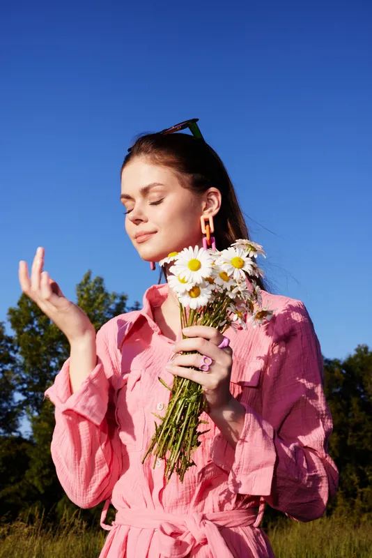 Young woman in a pink dress holding daisies, enjoying a sunny day outdoors, expressing joy and tranquility in a vibrant natural setting
