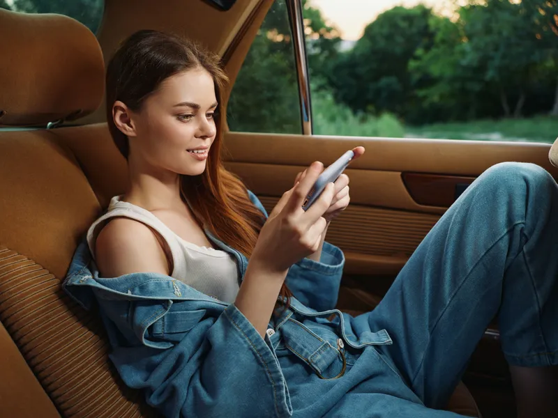 Girl enjoying her phone in a denim outfit inside a vintage car during golden hour, showcasing casual fashion and relaxed emotions.