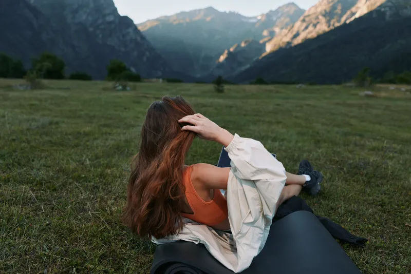 A woman sits in a meadow, leaning back on a small vehicle tire and gazing toward distant mountains. She wears a light jacket and relaxed expression, enjoying the outdoors and the sunlit landscape.