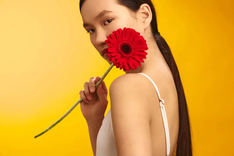asian woman in a light top smiles softly while holding a vivid red flower near her face, set against a sunny yellow background that creates a warm, inviting mood.