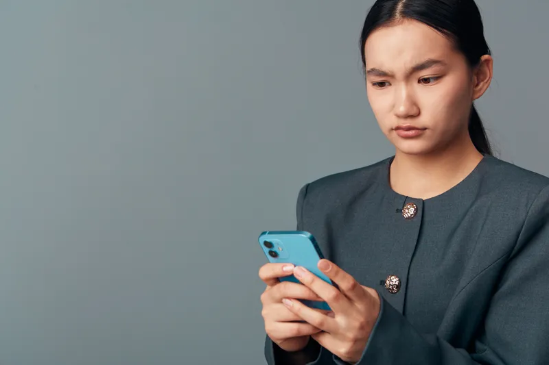 Woman with focused expression holding blue smartphone looking at screen with concern and concentration against plain gray background. Casual technology use and communication concept.