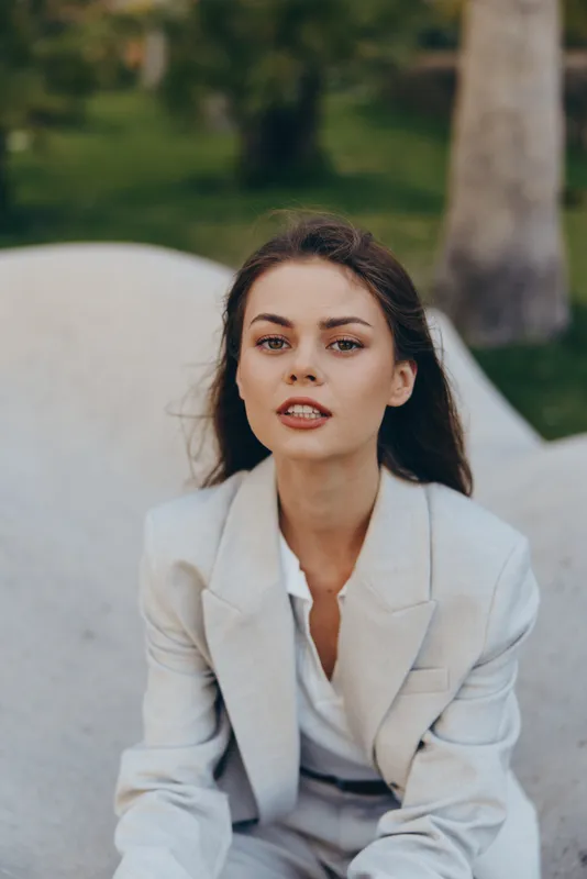 Woman in light grey business suit sitting outdoors with confident expression, natural background, professional business portrait with soft color palette