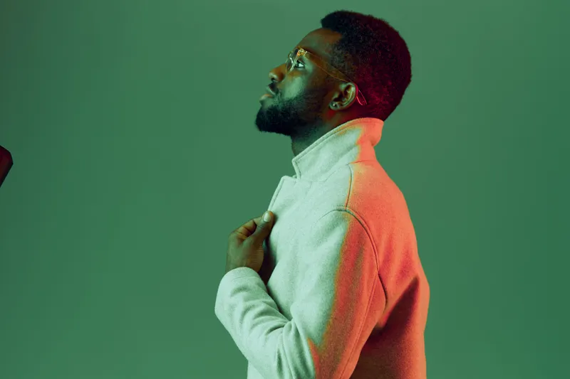 A profile of a Black male model wearing a light gray sweater against a green backdrop, exuding confidence and contemplation