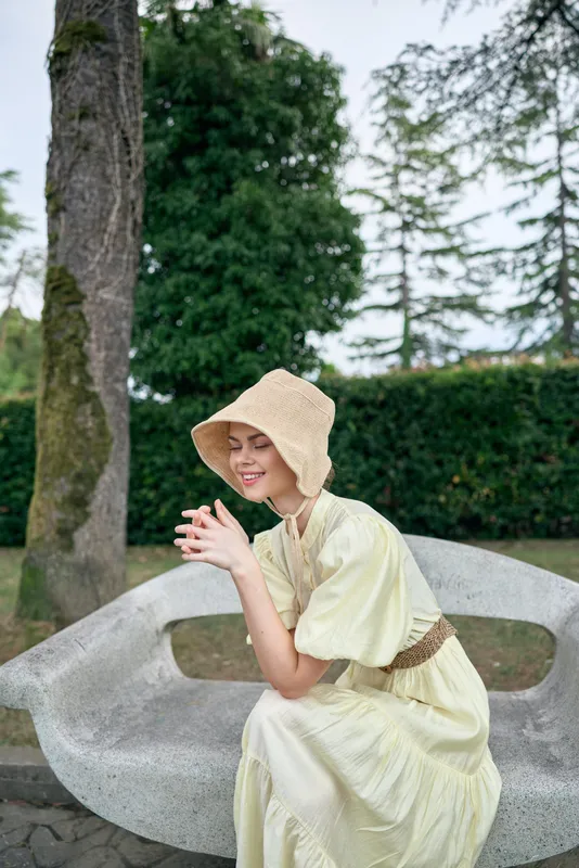Cheerful woman sits on a curved bench in a park, wearing a light yellow dress and a wide straw hat, smiling softly under trees, enjoying a warm sunny day in a peaceful outdoor setting