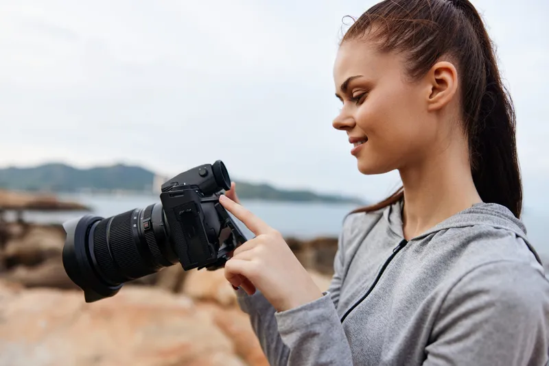 Young woman in a gray hoodie photographing scenic landscape with a DSLR camera at the beach during sunset, capturing moments and emotions