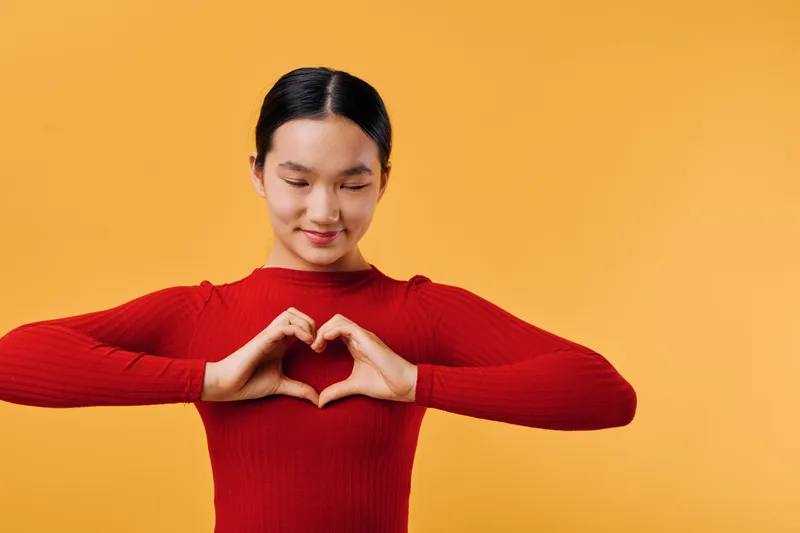Young woman in red sweater making heart shape with hands, smiling with closed eyes on orange background. Care, love and positivity gesture for emotion and connection.
