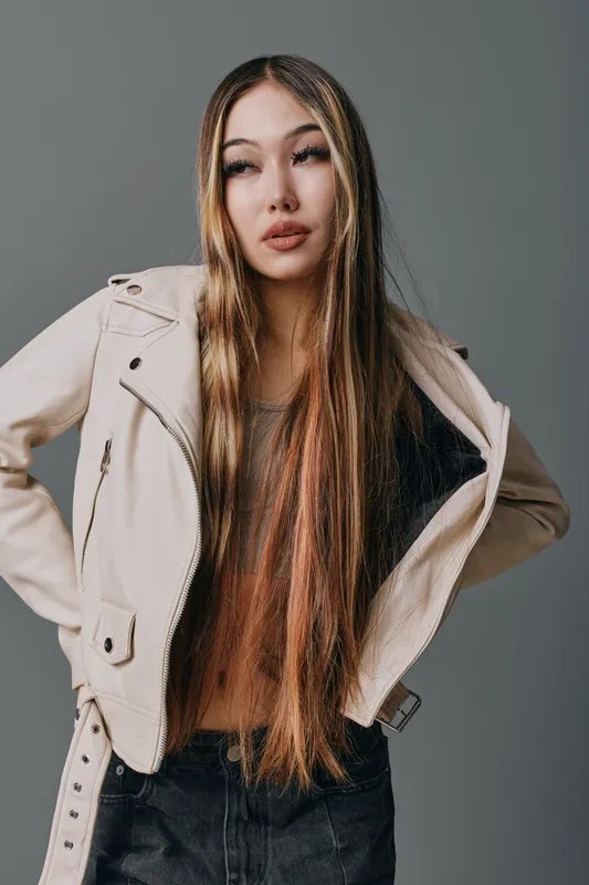 Young woman with long straight hair wearing beige jacket and dark pants posing with hands on hips against plain grey background in studio portrait.