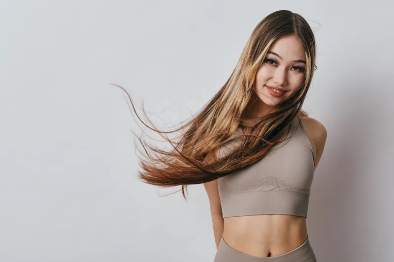 Young woman with long hair wearing beige sportswear smiling and posing on grey background. Fitness, beauty, casual style and healthy lifestyle concept with natural light.