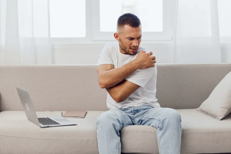 A man experiencing shoulder pain while seated on a couch, showcasing a contemporary and minimalistic living space. Focused on wellness and self-care in modern life.