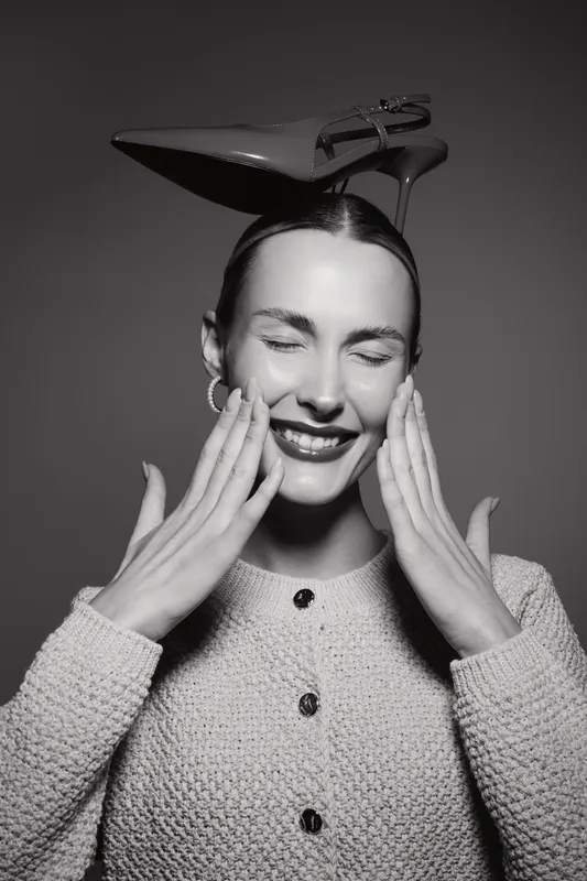 Smiling Caucasian woman with light skin wearing high heel shoe on head touches face with closed eyes in studio portrait.