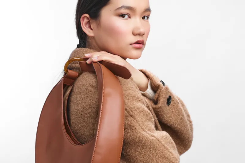 Asian woman wearing a tan coat stands with a brown bag over her shoulder in a minimal studio setting, exuding style, poise, and modern confidence.