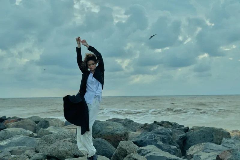 Young adult woman on rocky shore on windy day in pale dress and black jacket dancing by the sea under moody clouds travel