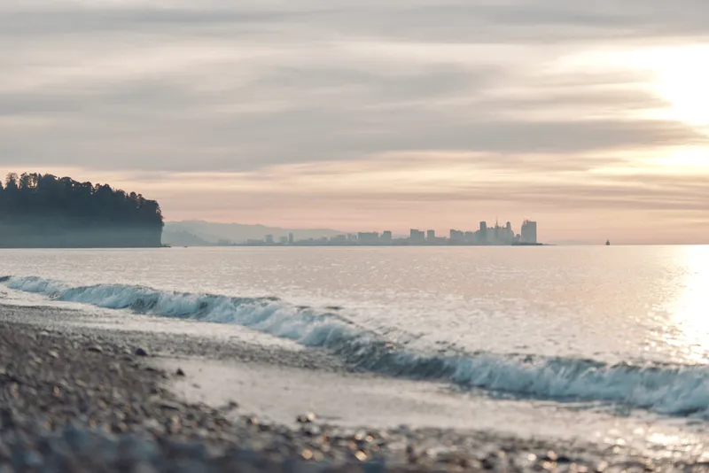 Beach and ocean scenery with gentle waves along a pebbled shore, sunset sky, distant city skyline on the horizon, calm water and tranquil coastline at dusk.