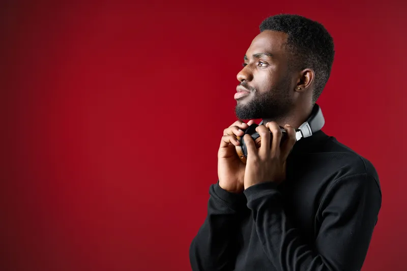 Stylish young Black man wearing headphones, gazing thoughtfully against a vibrant red background, perfect for music and technology themes