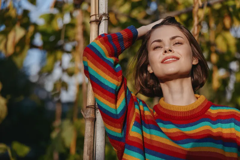 Woman portrait smile sweater outdoors sunlight autumn fashion relaxation, young woman in colorful striped jumper posing by a tree in warm natural light, cozy casual lifestyle and joy.