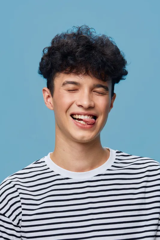 Smiling man with curly hair wearing a striped shirt playfully winking and sticking out his tongue against a solid blue background, showing fun and lively emotion.