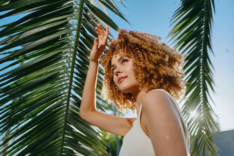 young woman with curly hair standing near green palm leaves under bright sunlight on clear blue sky background, wearing white sleeveless top, enjoying warm weather