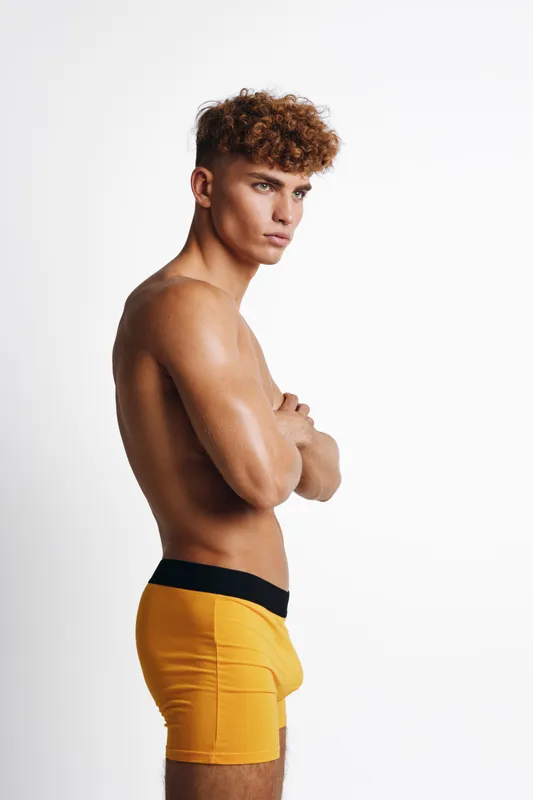 Young man with curly hair posing side view shirtless wearing yellow boxer shorts against white background studio isolated. Male model with muscular body serious facial expression and crossed arms.