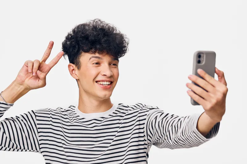 Young man taking selfie with smartphone, smiling happily and showing peace sign, wearing striped shirt against plain white background in casual style.