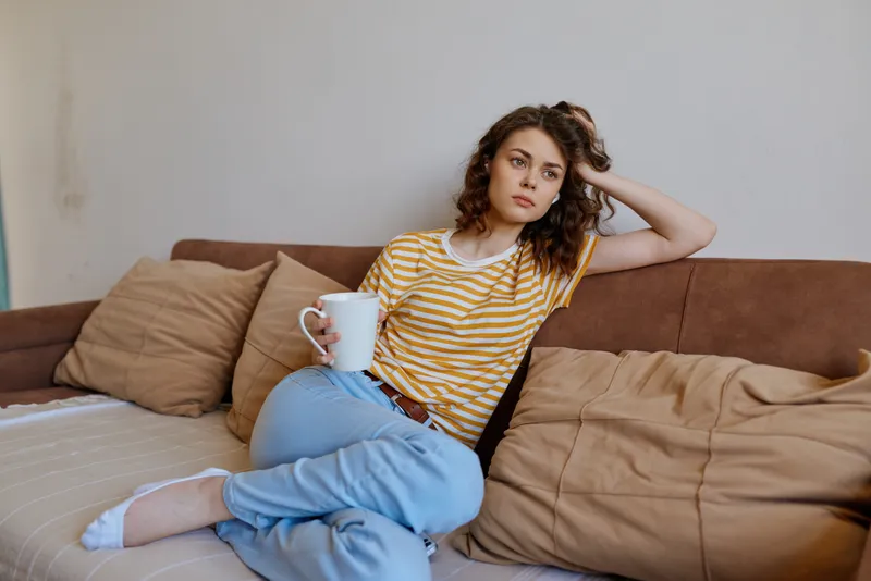 Woman lounging on a couch at home wearing a striped yellow shirt and jeans holding a mug relaxed mood casual living room setting with soft cushions natural daylight streaming in