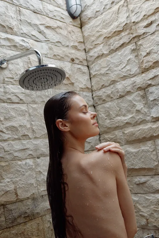 Naked woman enjoying outdoor shower with natural stone walls, wet hair, and relaxation, capturing peaceful moment in bright natural light.