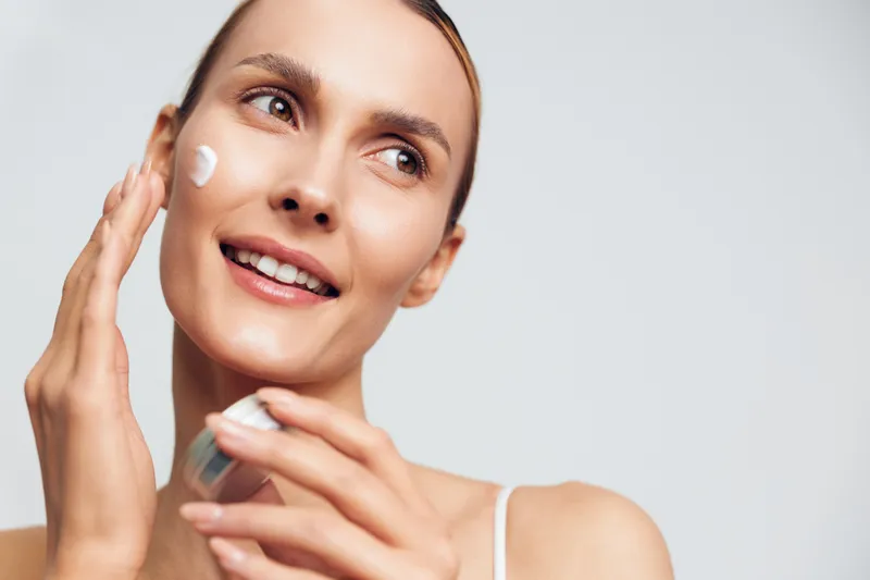 Smiling woman applying face cream with fingers, clear skin, natural light, skincare routine, beauty concept, closeup portrait, soft expression, neutral background.