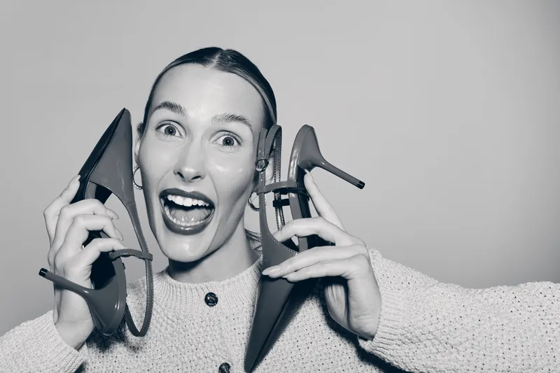 Happy white woman with light skin holds high heel shoes near face, smiling with wide open mouth. Studio portrait in black and white with joyful expression and casual sweater.
