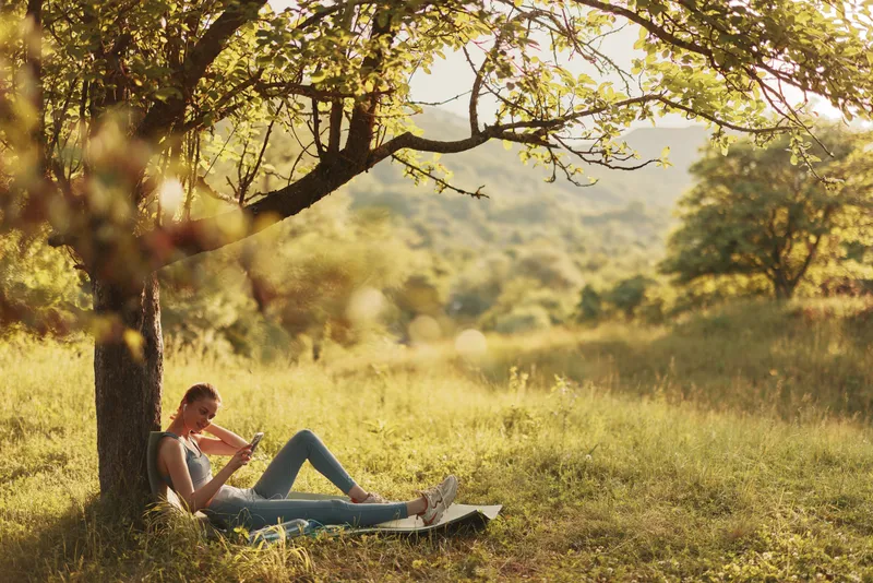 A young woman of European descent relaxing peacefully under a tree in a serene natural setting, enjoying the sunlight in casual attire, reflecting tranquility