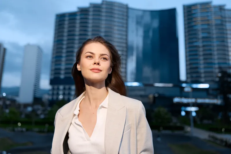 Confident woman in business attire against a modern city skyline, embodying professionalism and ambition during twilight hours.