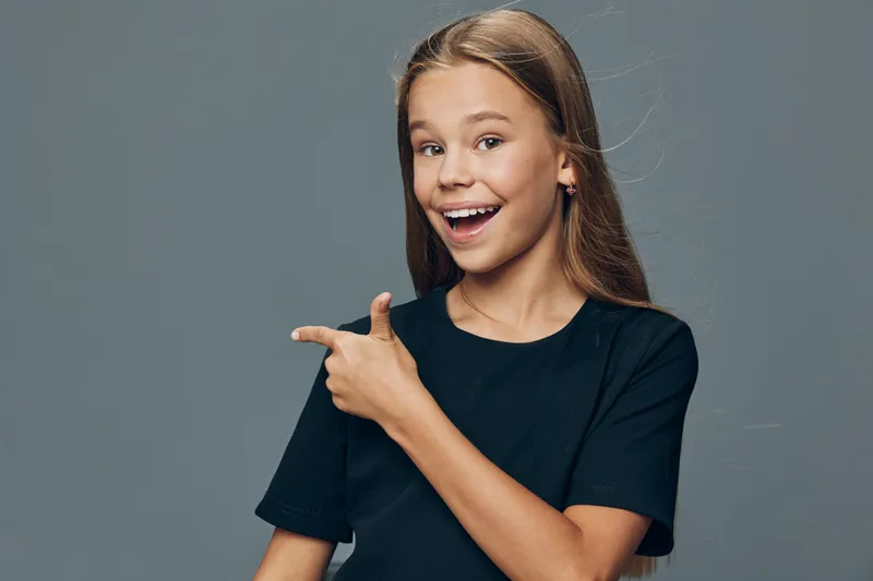 Girl child with long hair smiling and pointing to side against gray background. Happy young female wearing black shirt showing direction with finger gesture and cheerful expression.