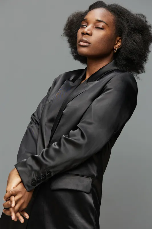 Confident young woman in black satin suit with natural curls posing against gray background, exuding empowerment and elegance