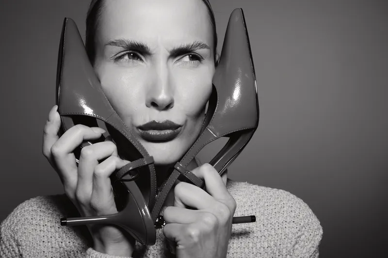 Serious Caucasian woman with pale skin holds high heel shoes near face in black and white portrait. Concept of fashion, style, and beauty.
