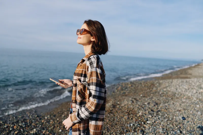 Woman on a pebble beach wearing a plaid shirt and sunglasses, holding a phone, enjoying the seaside breeze, distant waves and bright sky above.