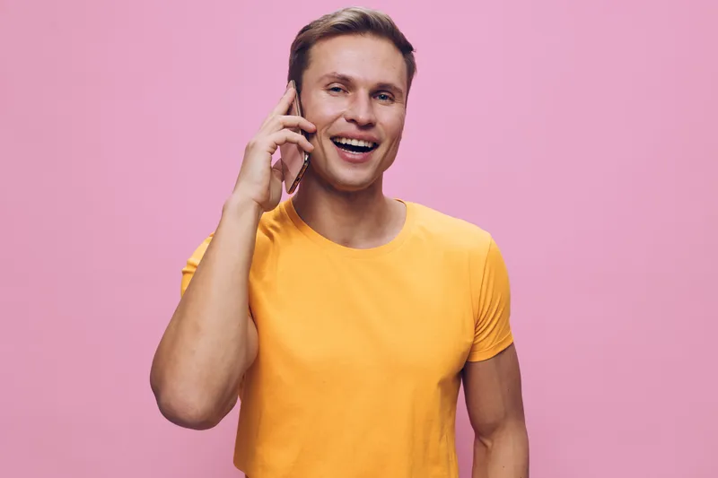 Smiling man talking on mobile phone has cheerful expression, wearing casual yellow t-shirt, standing against pink background. Studio portrait showcasing happiness, communication and positive emotions 