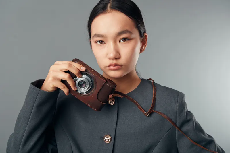 Young woman holding vintage camera looking at camera in studio with gray background, wearing dark blazer with buttons, serious expression, portrait of photographer or model